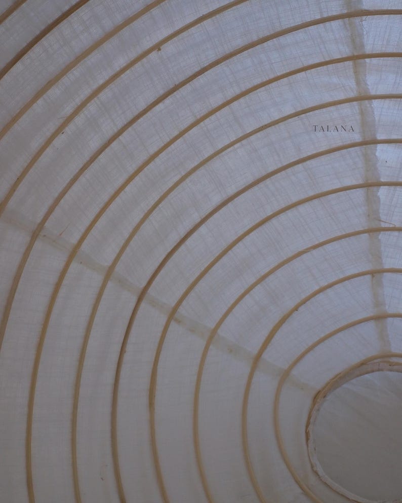 May include: A close-up view of a white, circular object with a series of curved, light brown wooden supports. The supports create a layered, concentric design. The word "TALANA" is printed in the upper right corner.