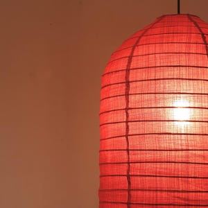 May include: A red paper lantern with a cylindrical shape, suspended from a black cord. The lantern's surface is divided into horizontal sections by darker lines, and the light source inside glows brightly.
