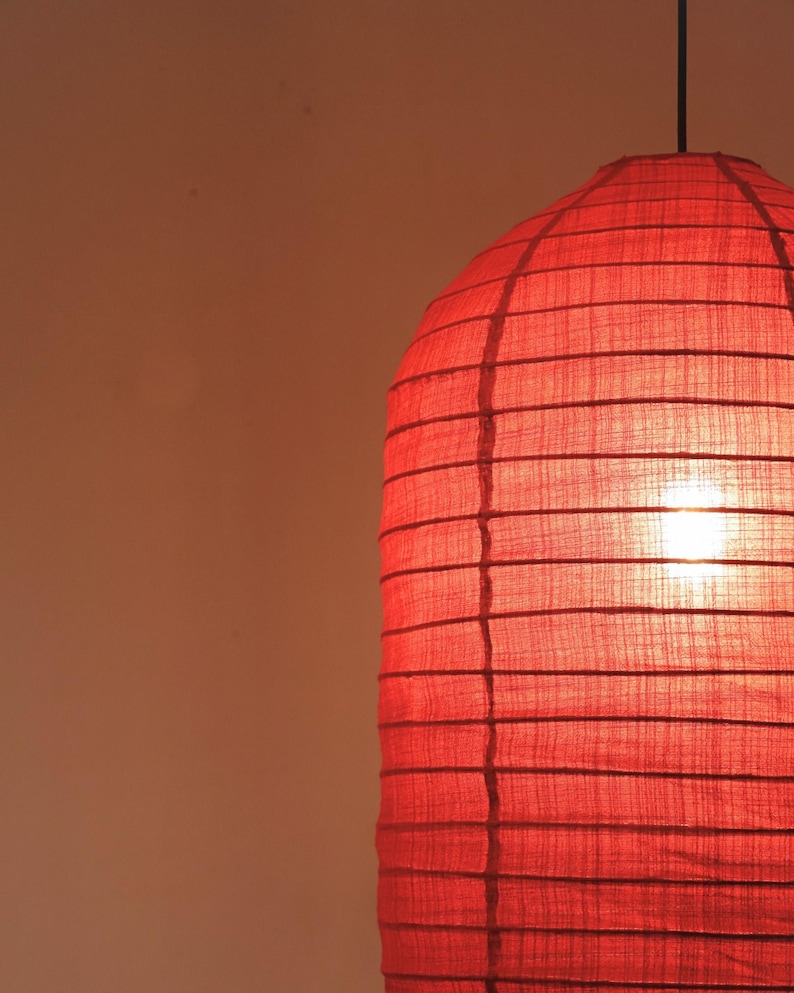 May include: A red paper lantern with a cylindrical shape, suspended from a black cord. The lantern's surface is divided into horizontal sections by darker lines, and the light source inside glows brightly.