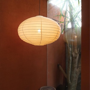 May include: A round, off-white paper pendant lamp with a ribbed texture, suspended by a thin black cord. The lamp emits a soft, warm glow, highlighting the horizontal wire frame structure visible through the translucent paper material.