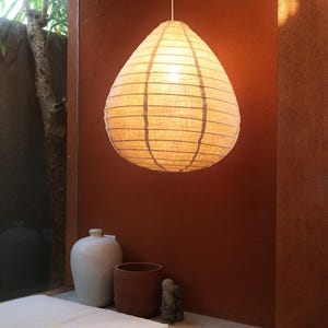 May include: A beige, teardrop-shaped pendant light hangs from a thin cord. The light is made of a textured, semi-transparent material, with vertical dark lines. The background features a terracotta-coloured wall and a glimpse of greenery.