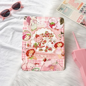 May include: A rectangular tablet case featuring a pink plaid background with illustrations of Strawberry Shortcake and Hello Kitty characters, strawberries, and cats. The text Strawberry Shortcake and Hello Kitty is printed in the center.