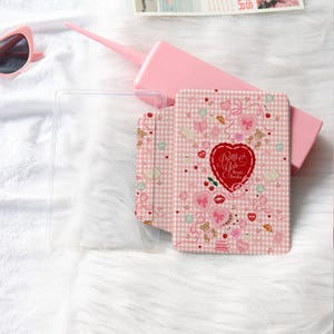Può includere: Una custodia per tablet a quadretti rosa e bianchi con la scritta "Pretty Girls Read Books" in un design a forma di cuore. La custodia presenta illustrazioni di cuori, fiocchi e orsetti. Sono visibili anche una custodia trasparente per tablet e occhiali da sole rosa.