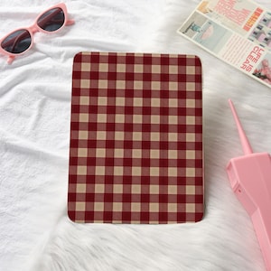 Op de afbeelding: Een rechthoekig item met een rood en beige ruitpatroon. Het item ligt op een witte, zachte ondergrond. Roze cat-eye zonnebril en een roze object zijn op de achtergrond.