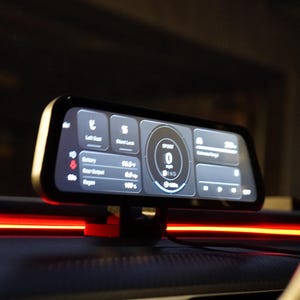 Può includere: Un display digitale rettangolare montato sul cruscotto di un veicolo, con una cornice nera e uno schermo luminoso con testo e icone bianche che mostrano velocità, stato della batteria e impostazioni del veicolo su sfondo scuro con luce rossa.