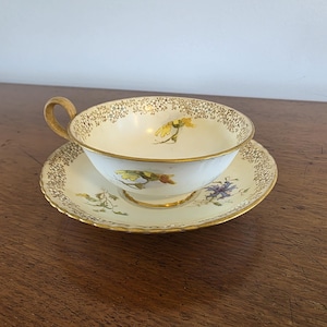 May include: A cream-coloured porcelain teacup and saucer set featuring delicate floral illustrations in yellow, green, and purple. The rim of both the cup and saucer is decorated with a fine gold filigree pattern and a solid gold edge trim.