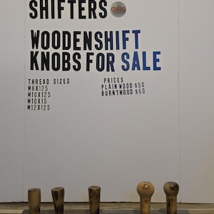 May include: Wooden shift knobs for sale. The sign reads "GREATGRAIN SHIFTERS WOODENSHIFT KNOBS FOR SALE". The knobs are made of wood and come in various shapes and finishes. Thread sizes include M8x125, M10x125, M10x15, and M12x125.