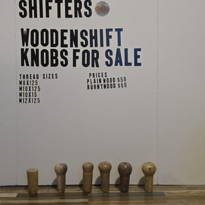 May include: A collection of wooden shift knobs in various shapes and shades of brown. The sign above reads "GREATGRAIN SHIFTERS WOODENSHIFT KNOBS FOR SALE" with thread sizes and prices listed. The knobs are displayed on a wooden floor.