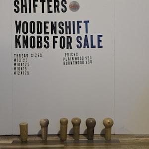 May include: A collection of wooden shift knobs in various shapes and shades of brown. The sign above reads "GREATGRAIN SHIFTERS WOODENSHIFT KNOBS FOR SALE" with thread sizes and prices listed. The knobs are displayed on a wooden floor.