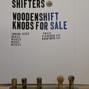 May include: Wooden shift knobs for sale. The sign reads "GREATGRAIN SHIFTERS WOODENSHIFT KNOBS FOR SALE." The knobs are made of wood and are various shapes and sizes. The thread sizes are M8X125, M10X125, M10X15, and M12X125.