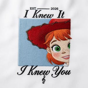 Op de afbeelding: Een witte stof met een geborduurd ontwerp van een personage met oranje haar en een rode hoed, tegen een lichtblauwe vierkante achtergrond. De tekst luidt EST 2026 I Knew It I Knew You, met een vioolsleutel onder het laatste woord.