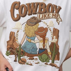 May include: A white t-shirt featuring an embroidered desert scene with two figures wearing cowboy hats, one with blonde hair and one with a red braid. The text reads COWBOY LIKE ME and I KNEW IT I KNEW YOU, surrounded by cacti, rocks, and a small horse.