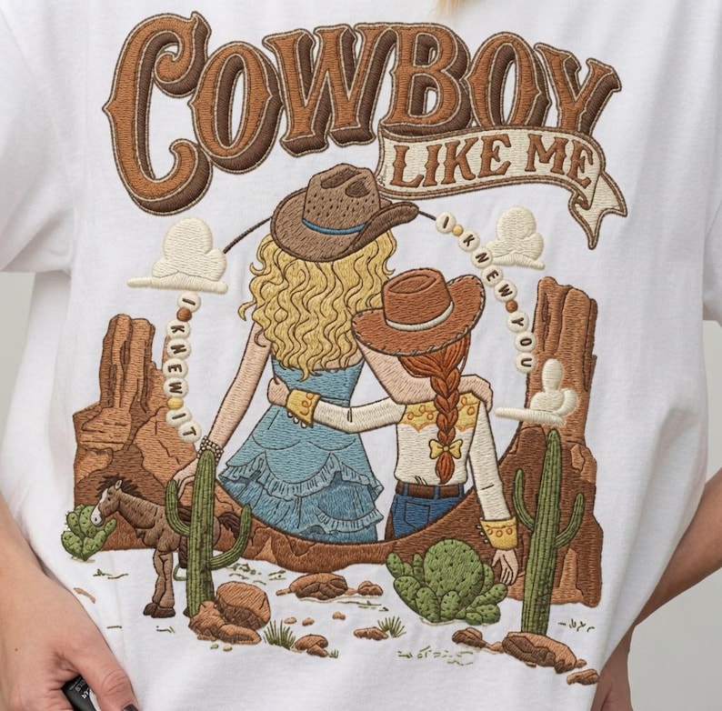 May include: A white t-shirt featuring an embroidered desert scene with two figures wearing cowboy hats, one with blonde hair and one with a red braid. The text reads COWBOY LIKE ME and I KNEW IT I KNEW YOU, surrounded by cacti, rocks, and a small horse.