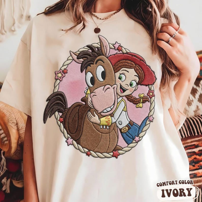 May include: An ivory t-shirt featuring an embroidered circular design of a brown horse being hugged by a character with red hair, a red hat, and a yellow shirt. The circular frame is made of a rope pattern with small pink and red stars.