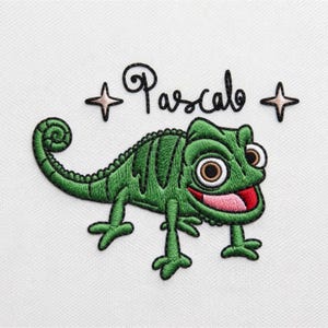 Op de afbeelding: Een geborduurde patch op een witte stoffen achtergrond met een groene kameleon met een gekrulde staart, grote ogen en een open mond die een roze tong laat zien. De naam Pascal is in zwarte cursieve letters erboven geborduurd, geflankeerd door twee kleine sterren.