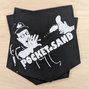 May include: A black fabric patch featuring a white screen-printed graphic of a person wearing glasses and a hat, throwing sand with one hand. The text POCKET SAND is printed in bold white capital letters across the lower portion of the design.