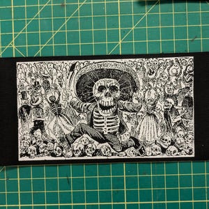 May include: Black and white patch featuring a Day of the Dead skeleton in a sombrero, surrounded by other skeletons. The patch has a rectangular shape and is made of fabric. The design is detailed and intricate.