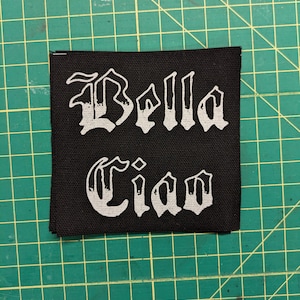 May include: Black square patch with the words "Bella Ciao" embroidered in white gothic lettering. The patch is made of a woven fabric and is likely designed to be sewn onto clothing or accessories.