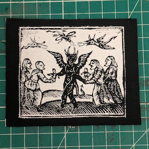 May include: A black and white patch featuring a detailed illustration of a horned demon with outstretched wings, surrounded by figures holding small dolls. Above the demon are three stylized flying creatures. The patch has a black border.