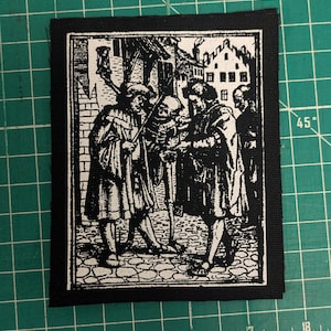 May include: Black and white patch featuring a historical illustration of several figures in period clothing, gathered around a skeleton. The scene is set in a town square with buildings in the background. The patch has a black border.
