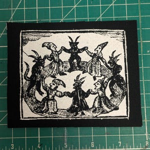 May include: Black and white patch featuring a circle of figures with horns and cloaks, dancing around a central horned figure. The patch is rectangular with a black border, and the design is printed on a white background.