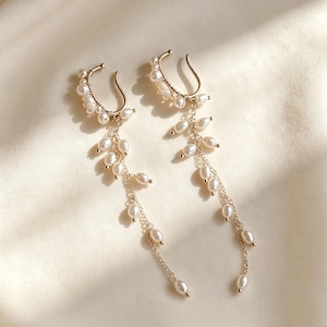 May include: A pair of gold-toned ear cuffs with dangling pearl accents. The ear cuffs curve around the ear, adorned with small pearls. Delicate chains with teardrop-shaped pearls hang from the cuffs, creating an elegant, flowing design.