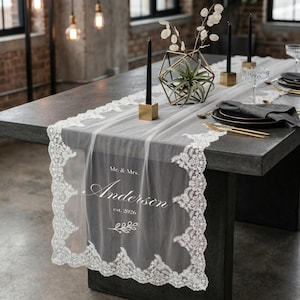 Peut inclure: Un chemin de table en dentelle blanche transparente avec un bord festonné, personnalisé avec le texte "Mr. & Mrs. Anderson est. 2026". La table est dressée avec des assiettes noires, des couverts dorés et des bougies noires dans des supports dorés.