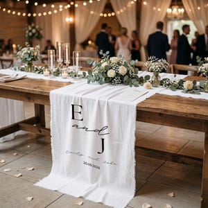 Op de afbeelding: Een witte stoffen tafelloper met gerafelde randen voorzien van de zwarte tekst E and J, Emily and John, en de datum 20.09.2026. De loper ligt op een lange houten tafel versierd met witte bloemstukken en glazen kandelaars.