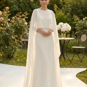 May include: A floor-length ivory gown featuring a sleeveless bodice with a round neckline and a sheer, flowing chiffon cape overlay that drapes from the shoulders to the floor, creating a soft, layered silhouette with a subtle train.