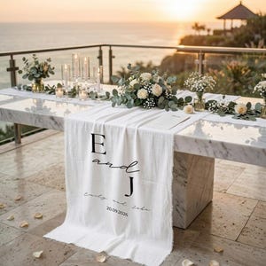 May include: A marble table is set for a wedding, with a white linen runner bearing the letters "E and J" and the date 20.09.2026. The table is adorned with floral arrangements, candles, and glassware, set against an ocean backdrop at sunset, with a gazebo in the distance.