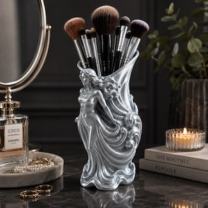 Puede incluir: Un soporte para brochas de maquillaje de resina en tono plateado con una figura femenina esculpida de cabello y vestimenta fluidos. El recipiente cilíndrico contiene una colección de varias brochas de maquillaje con cerdas oscuras.