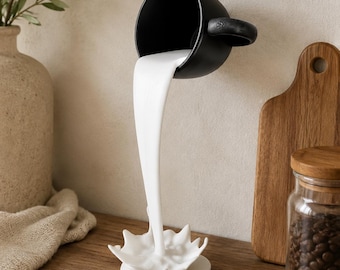 May include: A black ceramic mug seemingly pours a stream of white liquid into a white splash-shaped base. The mug is mounted on a wall. A wooden chopping board and a jar of coffee beans are in the background.