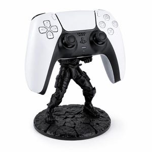 以下が含まれることがあります： 白と黒のPlayStation 5コントローラーが黒いフィギュアに支えられています。コントローラーは、黒いアクセントとボタンが付いた白い表面を持っています。フィギュアは、テクスチャ加工された黒い円形の台座に立っています。
