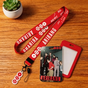 Puede incluir: Un cordón rojo con la palabra "ARIRANG" en blanco, junto con un portatarjetas a juego y un dije. El portatarjetas presenta una foto de un grupo de personas. El cordón también tiene los nombres de los miembros del grupo.