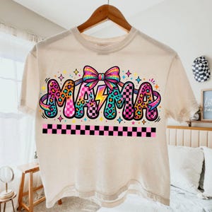 Puede incluir: Camiseta color crema con la palabra "MAMA" en un diseño retro colorido. La palabra está decorada con estampado de leopardo, patrones de cuadros y un rayo. Un lazo arcoíris y estrellas completan el diseño.