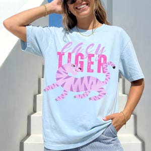 Puede incluir: Camiseta azul claro con un diseño de tigre rosa y el texto "easy tiger". El tigre tiene rayas moradas y una cara sonriente. La camiseta es de estilo casual.