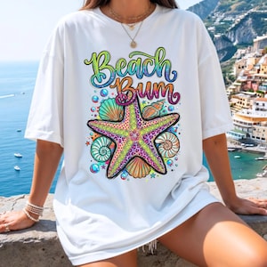 Puede incluir: Camiseta blanca con un gráfico colorido que dice "Beach Bum". El gráfico incluye una estrella de mar, conchas marinas y burbujas. La estrella de mar tiene un diseño de estampado de leopardo con colores rosa, amarillo y verde.