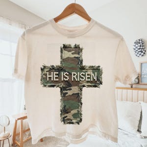 May include: Cream-colored t-shirt with a camouflage cross design. The cross is embellished with small white beads and the words "HE IS RISEN". The shirt is hanging on a wooden hanger.