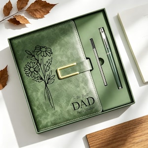 Könnte beinhalten: Grünes Leder-Notizbuch-Set mit Stift und Nachfüllung, präsentiert in einer Geschenkbox. Das Notizbuch hat ein Blumendesign und den Text "To the Best Dad" mit "Bella Daddy" darunter. Das Set enthält einen goldfarbenen Verschluss.