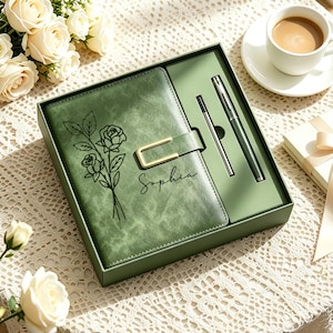 Può includere: Un set regalo con un diario in pelle verde con chiusura dorata, una penna e una matita. Il diario è impresso con un motivo floreale e il nome "Sophia". Una tazza di caffè e fiori sono sullo sfondo.
