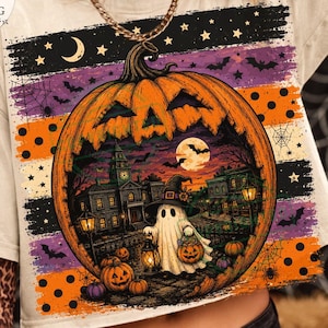 May include: A cream-colored t-shirt featuring a large graphic of a carved pumpkin framing a spooky scene with a ghost holding a lantern, a haunted house, bats, and stars. The background consists of horizontal stripes in black, purple, and orange.