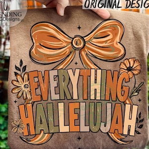 Op de afbeelding: Een bruin t-shirt met een grote grafische print van een strik en bloemen, voorzien van de tekst "EVERYTHING HALLELUJAH" in een dik, gelaagd lettertype. Het ontwerp gebruikt een kleurenpalet van oranje, beige en gedempt groen met steraccenten.