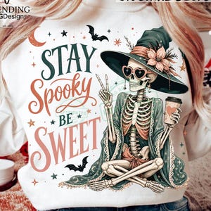 May include: A white sweatshirt featuring a graphic of a skeleton wearing a witch hat and heart-shaped sunglasses, holding a coffee cup and making a peace sign. The text reads STAY SPOOKY BE SWEET, surrounded by stars, a crescent moon, and bats.