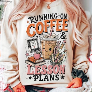 May include: A beige crewneck sweatshirt featuring a graphic print with the text RUNNING ON COFFEE & LESSON PLANS. The design includes a smiling iced coffee cup, a notebook labeled LEARN TEACH INSPIRE, a red apple, stacked books, and pencils.