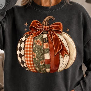 Könnte beinhalten: Ein anthrazitfarbenes Sweatshirt mit Rundhalsausschnitt und einer großen Kürbisgrafik, die aus vertikalen Paneelen mit verschiedenen Mustern wie schwarz-weißen Rauten, orangefarbenem Vichy-Karo, Blumenmuster und Streifen besteht, gekrönt von einer großen Samtschleife.