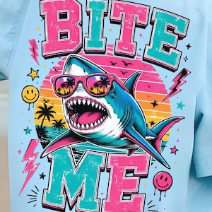 May include: Light blue t-shirt with a graphic of a shark wearing sunglasses, the text "BITE ME", and a sunset design. The shark is blue and white with pink accents. The shirt is a casual, beach-themed apparel item.