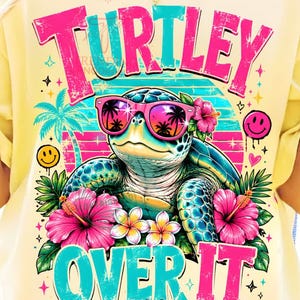 May include: Yellow t-shirt with a colorful graphic of a turtle wearing sunglasses and surrounded by flowers. The text reads "TURTLEY OVER IT" in pink and turquoise. The shirt has a relaxed fit.