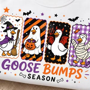Op de afbeelding: Een wit sweatshirt met ronde hals en een afbeelding van vier ganzen in Halloween-kostuums. De ganzen zijn verkleed als heks, spook, duivel en mummie. Onder de ganzen staat de tekst "GOOSE BUMPS SEASON" in paarse en oranje letters.