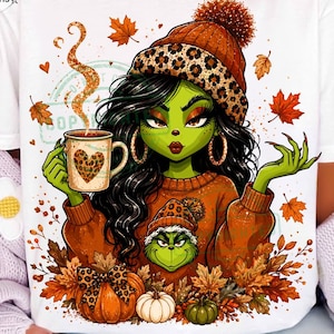 May include: A white t-shirt featuring a graphic of a green-skinned character with long black hair wearing a leopard print beanie and an orange sweater. The character holds a mug with a leopard print heart, surrounded by autumn leaves and pumpkins.