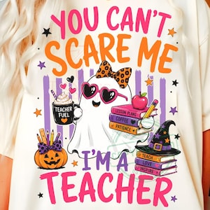Könnte beinhalten: Ein cremefarbenes T-Shirt mit einer Grafik eines weißen Geistes, der eine herzförmige Sonnenbrille und eine Leoparden-Schleife trägt. Der Text lautet "YOU CAN'T SCARE ME I'M A TEACHER", ergänzt durch Bücher, einen Kürbis und eine Tasse "TEACHER FUEL."